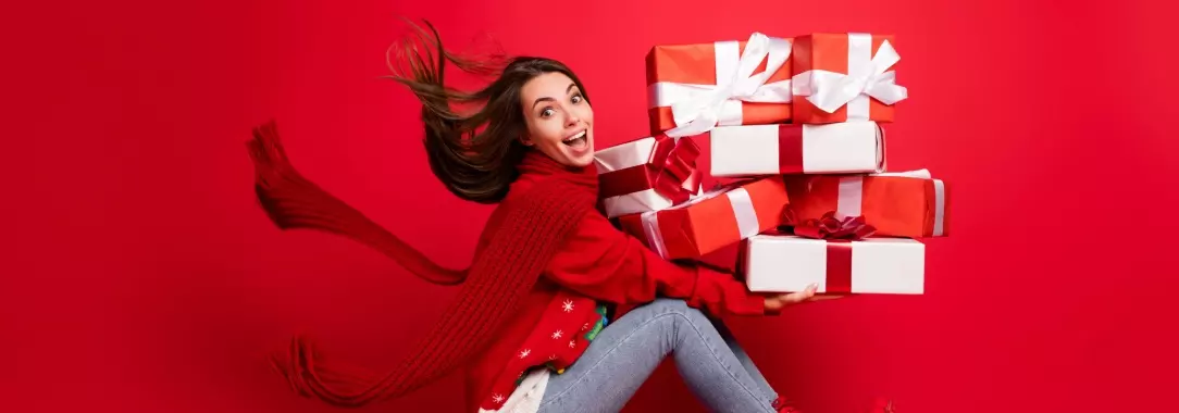 A photo of a festive woman sitting on a sleigh holding a pile of Christmas presents wrapped in red and white isolated on a red b