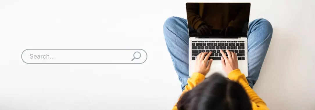 An image of a woman sitting cross-legged with a laptop on her lap and a search bar to her left on a light background