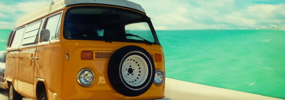 A retro looking photo of an old yellow VW van with the beach and ocean in the background 